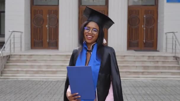 Uma tímida afro-americana graduada em um manto preto de um chapéu mestrado posa para a câmera. Um evento importante. Jovem especialista — Vídeo de Stock