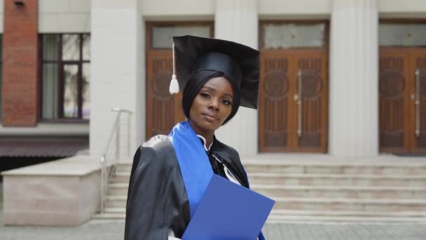 Donna afroamericana laureata in abito nero e cappello master quadrati in possesso di diploma su sfondo universitario. Un evento importante. Giovani specialisti — Video Stock