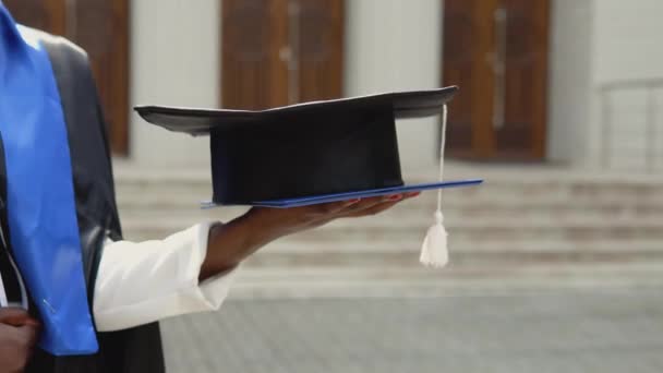 An African-American graduate woman in a black masters gown proudly and cheerfully holds a diploma and a square hat against the backdrop of the university. An important event. Close view of a hand — Stock Video