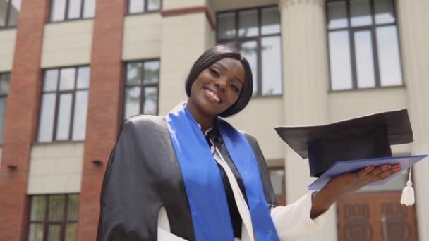 Een Afro-Amerikaanse afgestudeerde vrouw in een zwarte meesters jurk met een blauw lint op haar schouders trots en vrolijk houdt een diploma en een vierkante hoed tegen de achtergrond van de universiteit. — Stockvideo
