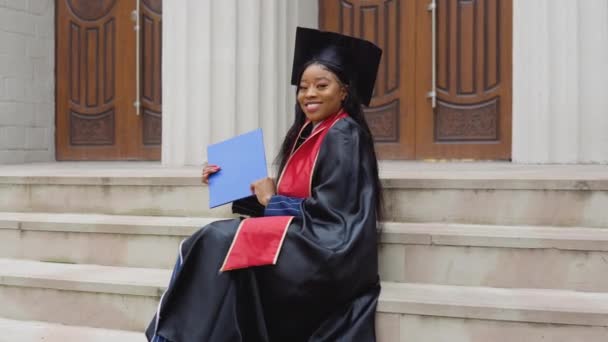 Femme afro-américaine diplômée en robe noire et un chapeau de maître avec un ruban rouge sur les épaules est assis sur les escaliers à l'entrée de l'université avec un diplôme d'enseignement supérieur en main. — Video