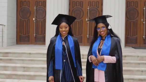 To afroamerikanske kvinner uteksamineres i klassiske masterdrakter og kapper står foran universitetsinngangen. Gratulerer med studentutvekslingsprogrammet. – stockvideo