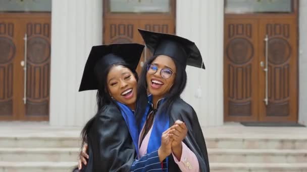 Dos mujeres afroamericanas graduadas en trajes y túnicas clásicas se paran abrazando frente a la entrada de la universidad. Graduados felices del programa de intercambio de estudiantes. — Vídeos de Stock