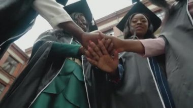 Üstün cübbeler ve kare şapkalarla üniversite ya da üniversite mezunları el ele tutuşarak takım ruhunu gösteriyor. Alt manzara. Avrupa 'da Afro-Amerikalı kız öğrenciler