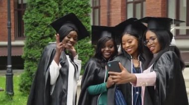 Mutlu üniversite ya da üniversite mezunları yüksek lisans elbiseleri ve kare şapkalar içinde canlıdır ve selfie çekerler. Üniversite binasının yanında Afro-Amerikalı kız öğrenciler duruyor.
