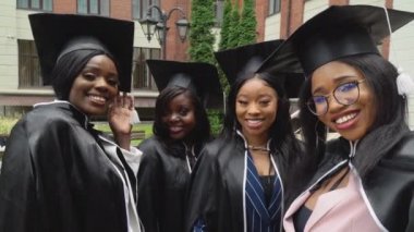 Mutlu üniversite ya da üniversite mezunları yüksek lisans elbiseleri ve kare şapkalar içinde canlıdır ve selfie çekerler. Üniversite binasının yanında Afro-Amerikalı kız öğrenciler duruyor.