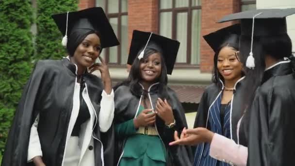 Glückliche Universitäts- oder Hochschulabsolventen in lebhaften Mänteln und quadratischen Hüten kommunizieren lebhaft und fröhlich. Afroamerikanische Studentinnen stehen in der Nähe des Universitätsgebäudes — Stockvideo