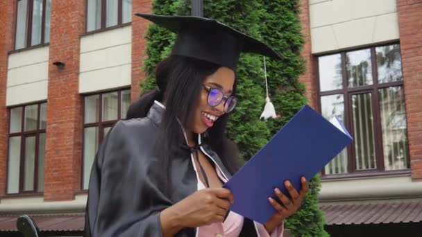 Eine afroamerikanische Absolventin in Mantel und Meistermütze sitzt mit einem Hochschuldiplom in der Hand im Hof der Universität. Ein lang erwarteter Erfolg — Stockvideo
