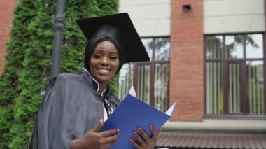 Bir Afro-Amerikalı kadın, bir manto ve bir master şapkası ile mezun olurken üniversitenin yanında otururken diplomasındaki notları mutlulukla okur. Başarın için gurur duy.