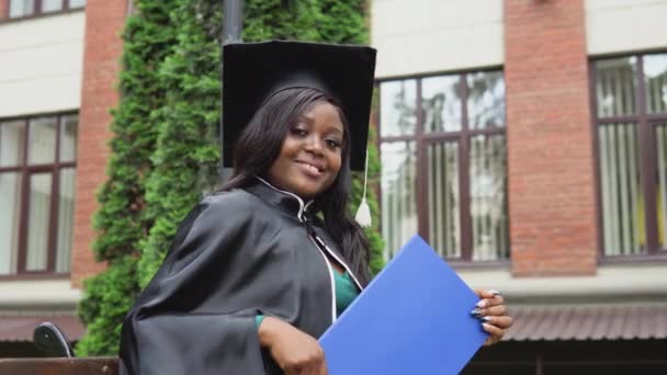 Una mujer afroamericana graduada en un manto y un sombrero de máster se sienta con un diploma de educación superior en sus manos. Una nueva etapa de la vida, inicio de una trayectoria profesional — Vídeo de stock