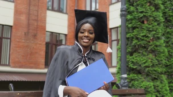 Mujer afroamericana graduada en un manto y un sombrero de máster sonriendo felizmente con un diploma en la mano mientras está sentada cerca de la universidad. — Vídeos de Stock