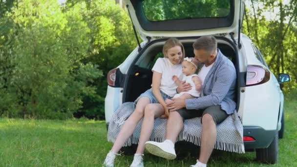 Familienfreizeit im Sommer. Papa, Mutter und Baby sitzen im Kofferraum eines weißen Familienautos auf einer Wiese vor den Toren der Stadt — Stockvideo