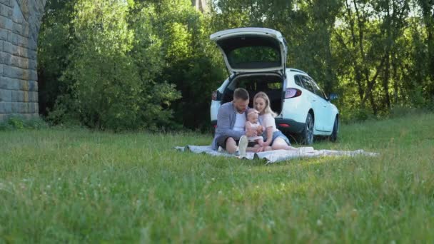 A boldog család családi autóval utazik. Piknik a városon kívül. Apa, anya és lánya egy szőnyegen ülnek a pázsit közepén. — Stock videók