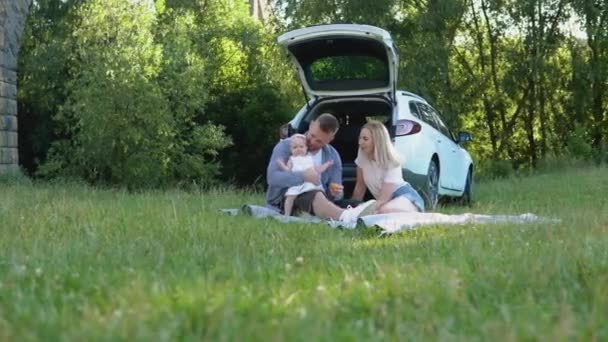 La familia feliz viaja en coche familiar. Picnic fuera de la ciudad. Padre, madre e hija están sentadas en una alfombra en medio del césped — Vídeos de Stock