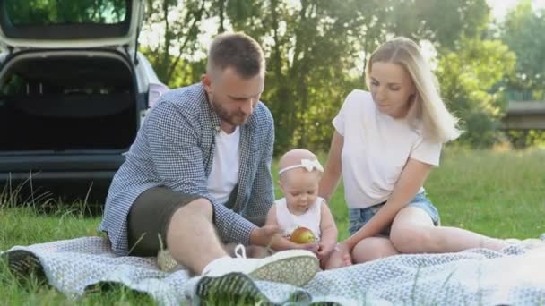Szczęśliwa rodzina podróżuje rodzinnym samochodem. Piknik z owocami poza miastem. Ojciec, matka i córka siedzą na dywanie na środku trawnika. — Wideo stockowe