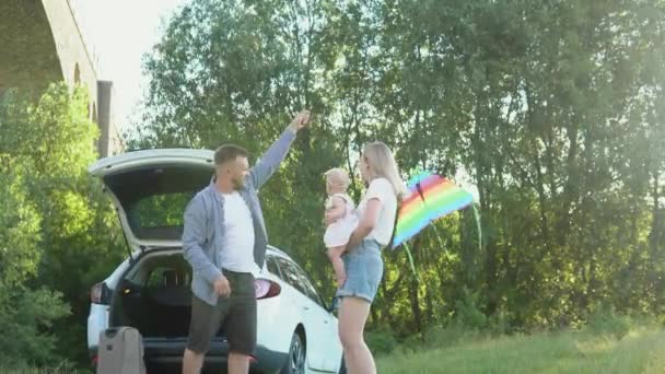 Een gelukkig gezin rust buiten de stad en reist met de auto. De vader lanceert een kleurrijke vlieger, en de moeder houdt de baby in haar armen. Een klein kind speelt met een vlieger — Stockvideo