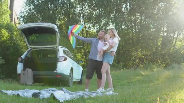 Una familia feliz está descansando fuera de la ciudad y viajando en coche. El padre lanza una cometa colorida, y la madre sostiene al bebé en sus brazos. Un niño pequeño juega con una cometa — Vídeos de Stock