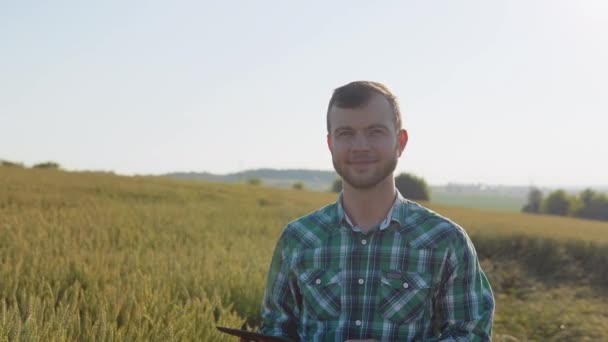 A young farmer agronomist with a beard stands in a field of wheat under a clear blue sky and holds documents in one hand and shows a thumb up with the other hand. Harvest in late summer — Stock Video