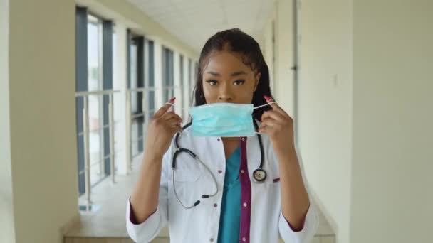 Joven mujer médica afroamericana en un traje azul y abrigo blanco lleva una mascarilla estéril desechable — Vídeos de Stock