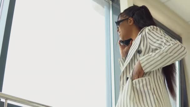 Гламурная афро-американская деловая женщина в черных очках и в белом полосатом костюме стоит у окна в коридоре офисного центра и эмоционально разговаривает по мобильному телефону — стоковое видео