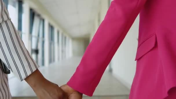 Dos glamurosas empresarias afroamericanas con traje blanco a rayas y traje rosa se toman de la mano y caminan por el pasillo del centro de oficinas frente a la cámara. Afro americano lesbianas pareja — Vídeos de Stock