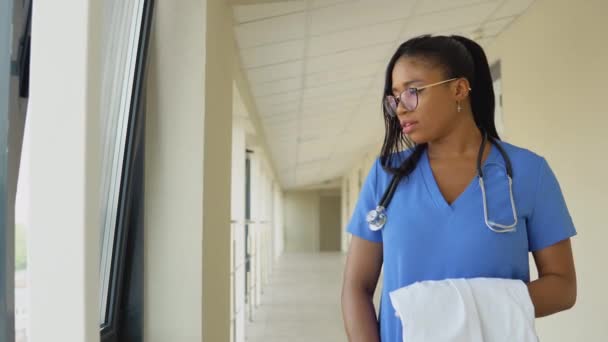 Egy fiatal afro-amerikai orvos nő kék öltönyben áll az ablaknál a klinika folyosóján fürdőruhában a kezében. — Stock videók