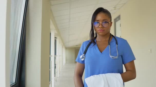 Jovem médica afro-americana de fato azul está à janela no corredor da clínica com um fato de banho nas mãos. — Vídeo de Stock