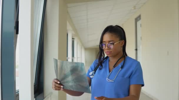Una giovane dottoressa afro-americana vestita di blu si trova nel corridoio della clinica ed esamina una radiografia di un torace adulto — Video Stock
