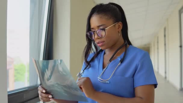 Uma jovem médica afro-americana em um terno azul fica no corredor da clínica e examina um raio-x de um peito de adultos. — Vídeo de Stock