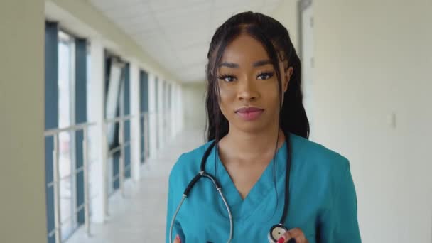 Una joven doctora afroamericana con traje azul y estetoscopio se encuentra en el pasillo de la clínica y mira con una sonrisa a la cámara — Vídeo de stock