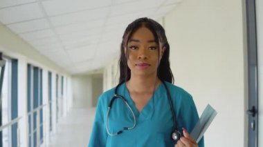 Mavi takım elbiseli genç bir Afrikalı Amerikalı doktor kliniğin koridorunda dikilip kameraya bakıyor. Doktorun elinde röntgen var.