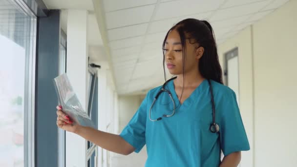 Una giovane dottoressa afro-americana vestita di blu si trova nel corridoio della clinica ed esamina una radiografia di un torace adulto — Video Stock