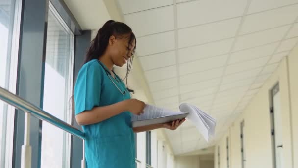 Jovem médica afro-americana em terno azul fica no corredor da clínica e vira documentos — Vídeo de Stock