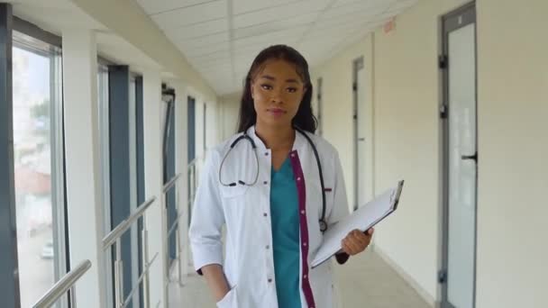 Young African American doctor woman in a blue suit and white coat walks along the corridor of the clinic with documents in her hand — Stock Video