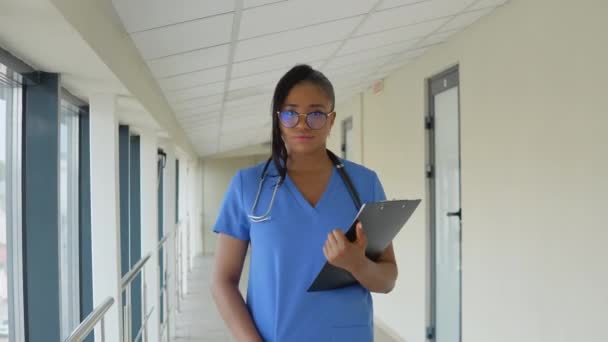 Giovane donna dottoressa afroamericana in abito blu cammina lungo il corridoio della clinica con i documenti in mano — Video Stock