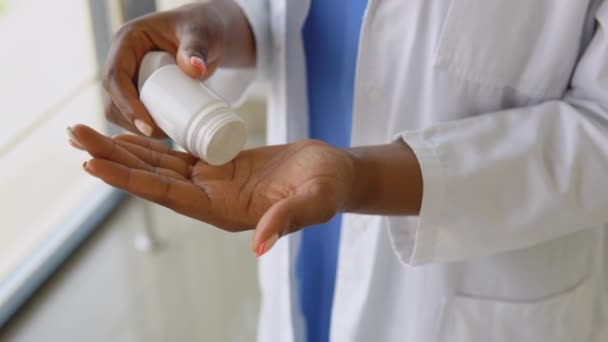 Mulher médica afro-americana em um terno azul e casaco branco derrama pílulas de um frasco na palma da mão. Visão de perto de mãos — Vídeo de Stock