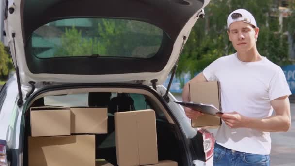 Traslado y entrega de productos manufacturados. Un empleado sostiene una caja en sus manos y se para junto a un coche lleno de paquetes — Vídeos de Stock