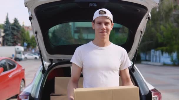 Bir çalışan elinde bir kutu tutuyor ve paketlerle dolu bir arabanın yanında duruyor. Üretilmiş malların nakliyesi ve teslimatı — Stok video