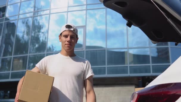 Traslado y entrega de productos manufacturados. Un empleado sostiene una caja en sus manos y se para junto a un coche lleno de paquetes — Vídeo de stock