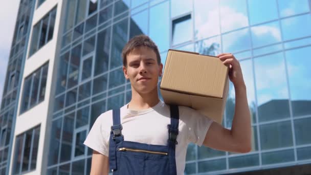 Un livreur tient une boîte sur son épaule, regarde la caméra, puis marche sur le côté. Livraison des marchandises au bureau — Video