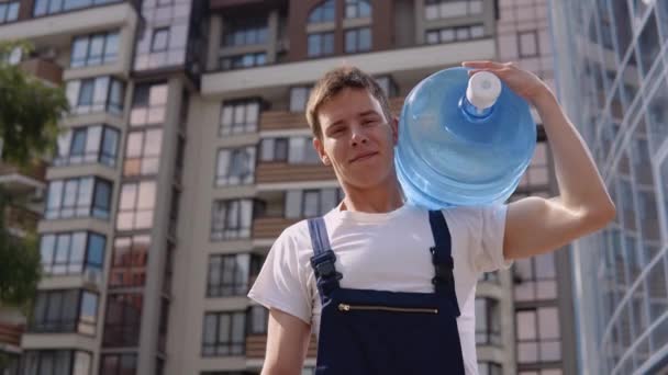 Курьер держит большую банку питьевой воды на плече на фоне современного высотного здания и смотрит в камеру. Доставка воды домой — стоковое видео