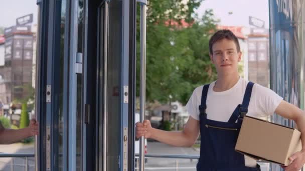 Ein Kurier in weißem T-Shirt und blauem Overall hält in der einen Hand eine Schachtel und öffnet mit der anderen die Glastür eines modernen Gebäudes. — Stockvideo