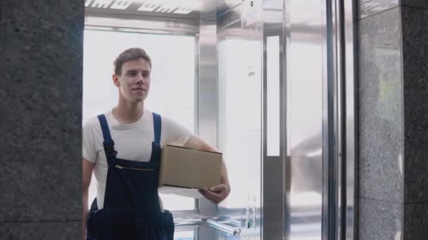 Um trabalhador de entrega em uma camiseta branca e macacão azul sai do elevador segurando uma caixa em sua mão. Entrega de mercadorias à estância aduaneira — Vídeo de Stock
