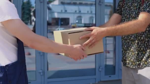 Un repartidor con una camiseta blanca y un mono azul entrega la caja a otro destinatario masculino cerca de la moderna puerta principal de cristal del rascacielos. Entrega de mercancías a la oficina — Vídeo de stock