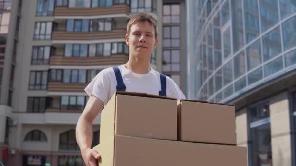 Leveringsmedewerker in een wit T-shirt en blauwe overall heeft drie boxen op de achtergrond van moderne hoogbouw. Exprespost — Stockvideo