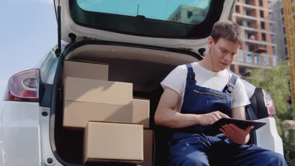 De koerier zit aan de rand van de open kofferbak, gevuld met dozen met goederen en vult documenten in — Stockvideo