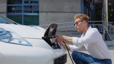 Genç bir kıvırcık adam elektrikli bir arabayı şarja bağlıyor.