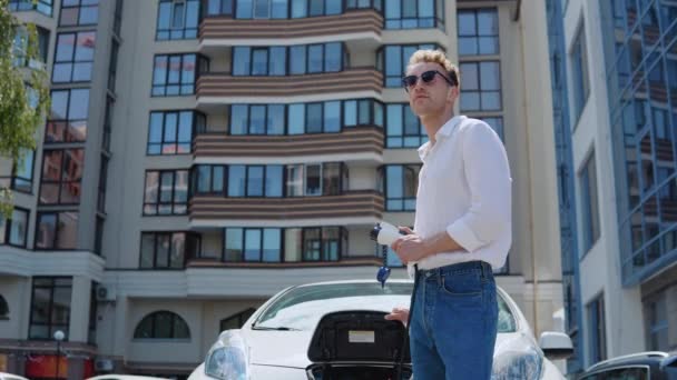 Een jonge krullende man in jeans en een wit shirt staat naast een elektrische auto met een oplader in zijn hand — Stockvideo