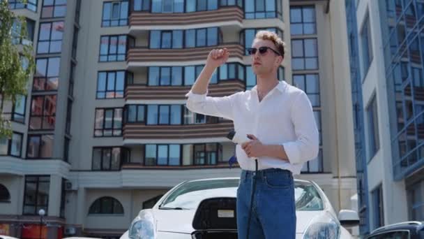 Snygg modern ung lockig man i jeans och en vit skjorta står nära en elbil med en laddare i handen på gården i ett bostadsområde — Stockvideo