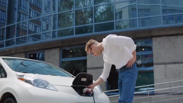 Stijlvolle moderne jonge krullende man in jeans en een wit shirt in de tuin van een wooncomplex ontkoppelt de elektrische auto van het opladen — Stockvideo